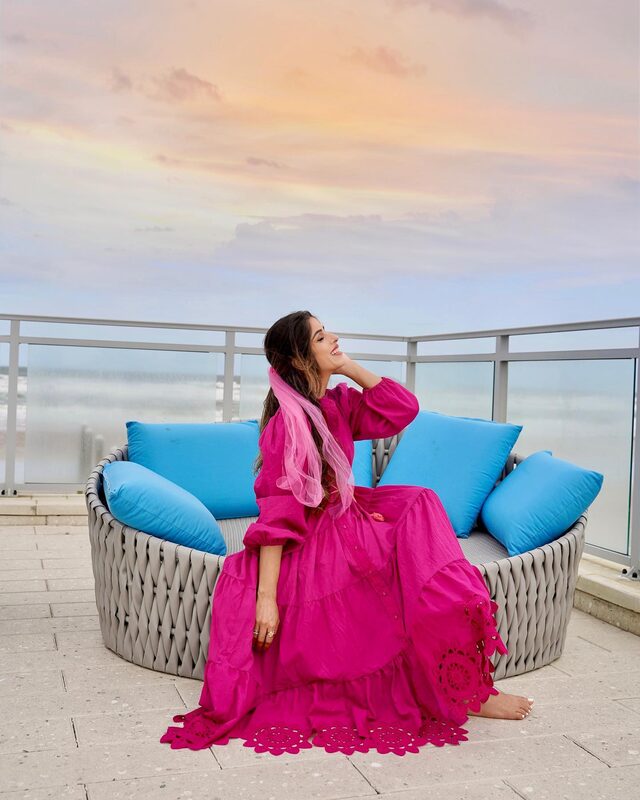 Roopsi Bansal in a magenta boho maxi dress from Chicwish with a sheer pink scarf, seated on an oceanfront balcony daybed with blue pillows at The Hard Rock Hotel Daytona Beach