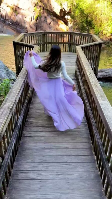 Roopsi Bansal in a white sweater and flowing lilac skirt from Chicwish, running across a wooden overlook deck above Tallulah Gorge with the rock face and river visible below