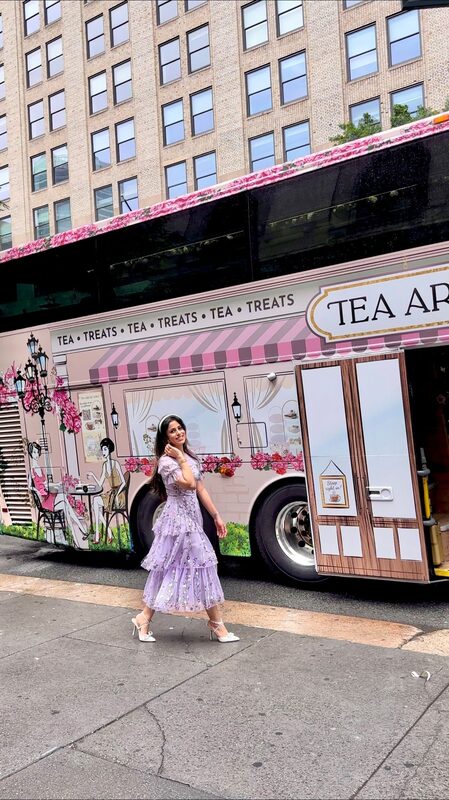 Roopsi Bansal in a lavender floral midi dress walking past the pink Tea Around Town NYC tour bus