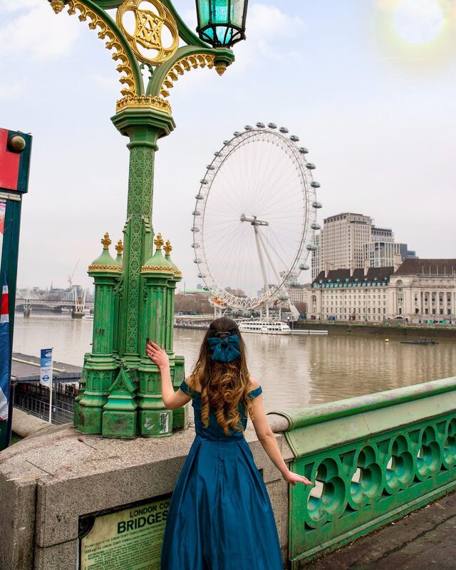 Roopsi Bansal from behind in a teal off-shoulder gown with a matching bow in her hair, standing on Westminster Bridge looking across the Thames at the London Eye