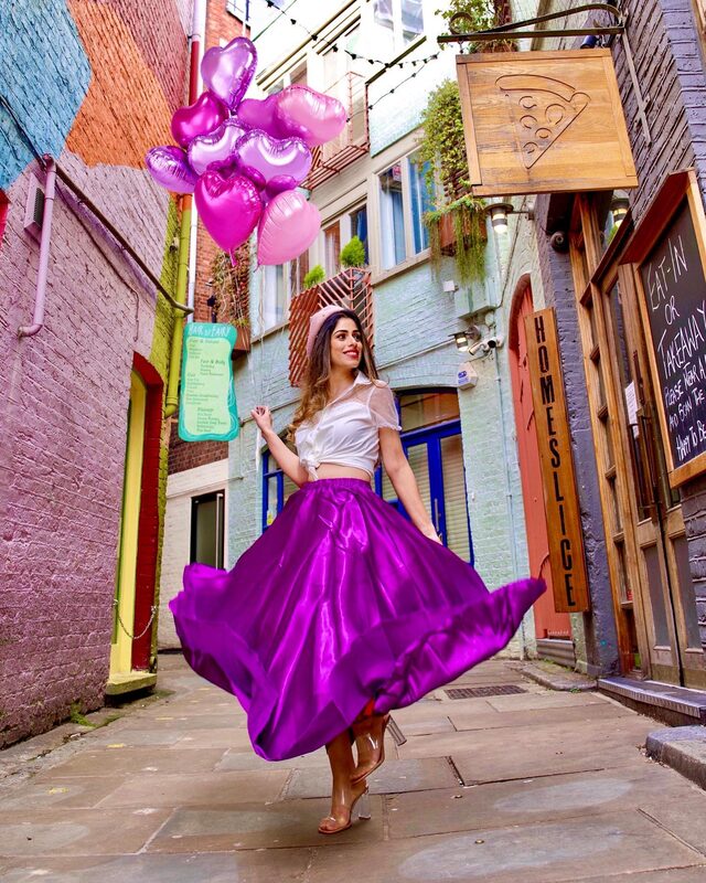 Roopsi Bansal twirling in a magenta ballgown skirt and white blouse, holding a bouquet of pink and purple heart-shaped balloons in the colourful alley at Neal's Yard, London
