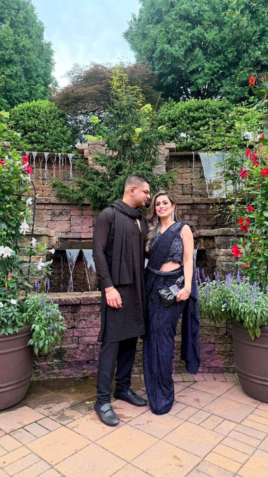 Roopsi Bansal in a navy sequined saree with her partner in a black kurta, standing in front of a stone garden fountain