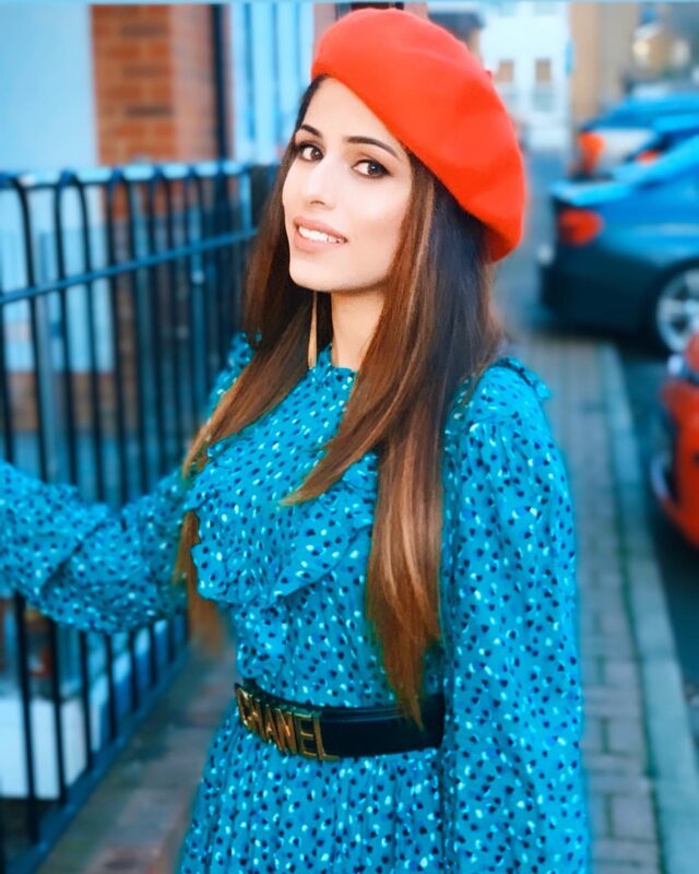Close-up shot wearing a green polka-dot dress and red beret, with a Chanel logo belt, standing by a black iron railing near a parked car