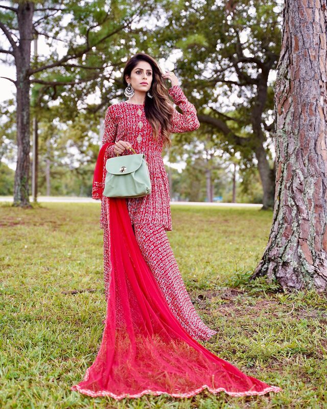 Roopsi Bansal in a red printed kurta set with a flowing sheer red dupatta and silver jhumka earrings, holding a mint green handbag under pine trees at Okeeheelee Park