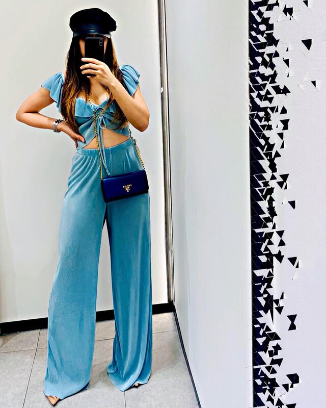 Mirror selfie wearing a light blue pleated tie-front crop top and matching wide-leg pants, a black baker-boy cap, and a blue Prada crossbody bag