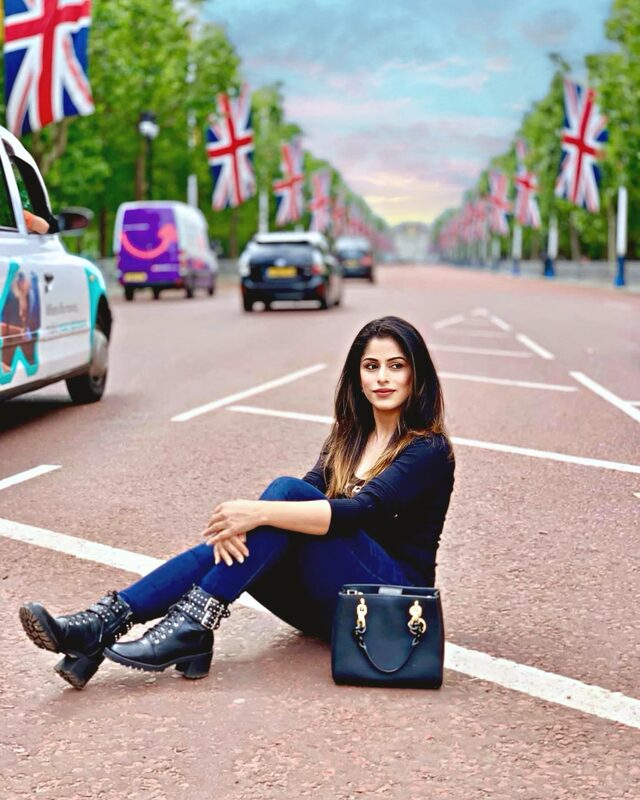 Sitting in the middle of a flag-lined avenue leading toward Buckingham Palace, wearing a navy top, jeans, and studded combat boots, with a black tote bag beside her