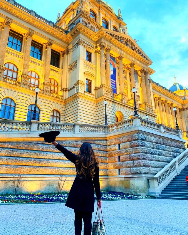 Standing before Prague's National Museum lit up gold at dusk, black coat and hat raised triumphantly in one hand, carrying a Burberry check tote