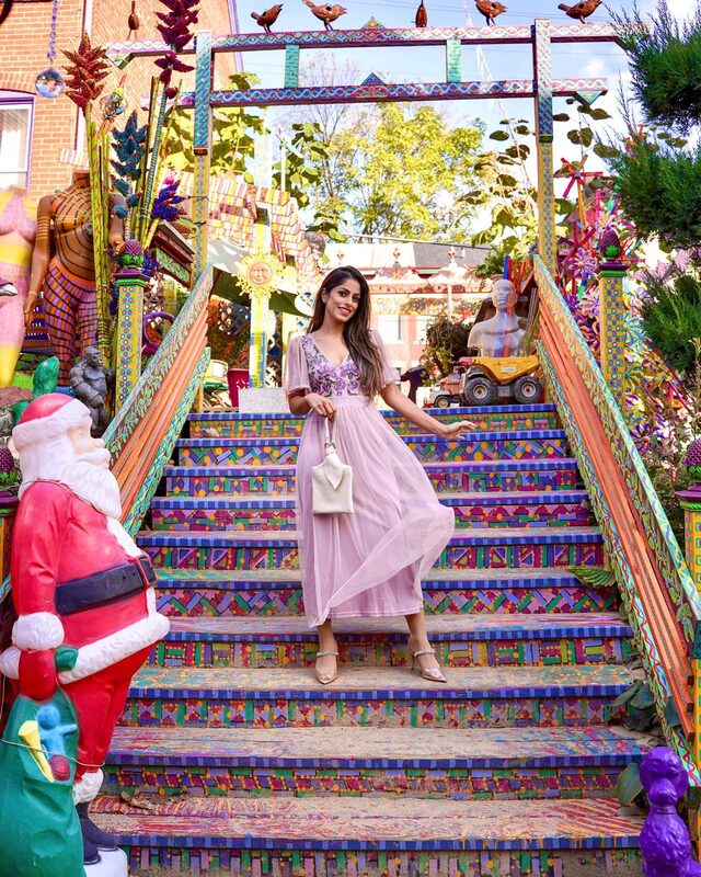 Roopsi Bansal in a lilac floral-embroidered dress from Frock and Frill, standing on a rainbow-painted staircase surrounded by folk-art sculptures and lawn ornaments at Randyland, Pittsburgh