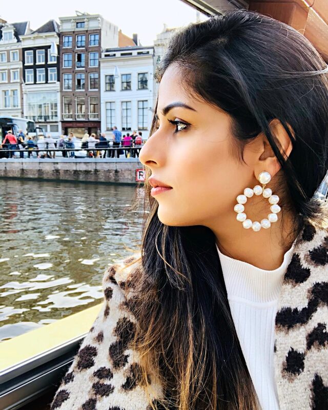 Close-up profile portrait on a canal boat in Amsterdam, wearing pearl hoop earrings and a leopard-print coat, with canal houses lining the water behind