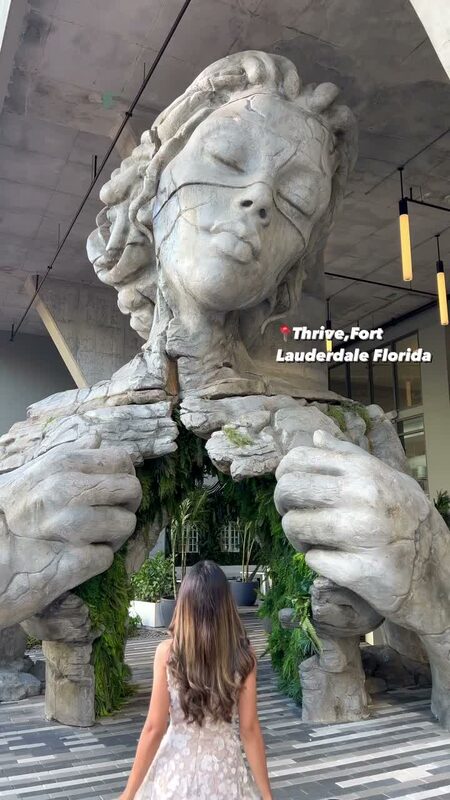 Roopsi Bansal in a white floral print dress from Dress the Population, standing beneath the towering stone Thrive sculpture, a monumental figure whose hands frame a garden passage, in Fort Lauderdale