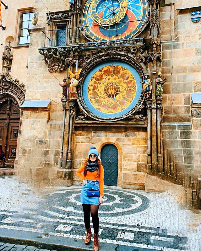 Standing beneath Prague's ornate Astronomical Clock, wearing an orange sweater, denim skirt, grey pom-pom beanie, and a blue Prada bag