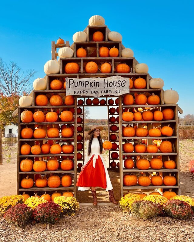 Roopsi Bansal in a white sweater and red pleated skirt holding a pumpkin in the doorway of the Pumpkin House at Happy Day Farm, New Jersey