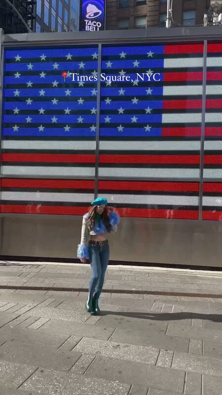 Roopsi Bansal in a teal beret and denim outfit standing in front of a giant American flag LED display in Times Square
