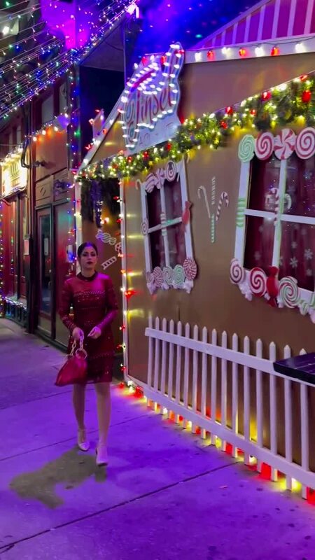 Roopsi Bansal in a red embellished dress walking past the candy-cane-lit gingerbread facade of Tinsel, a holiday pop-up bar in Philadelphia, at night