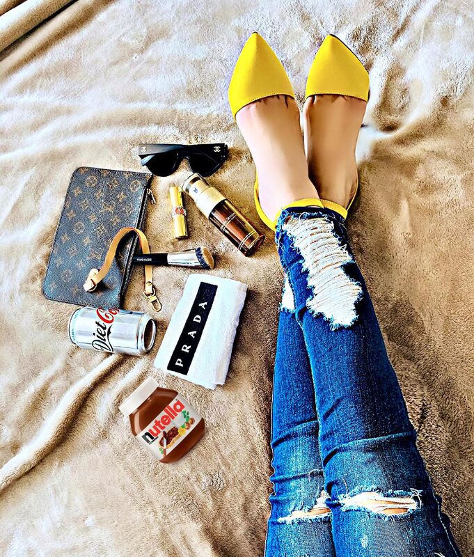 Flat-lay of legs in ripped jeans and yellow pointed flats stretched across a blanket, surrounded by a Louis Vuitton pouch, Chanel sunglasses, a gold perfume bottle, a makeup brush, a Prada cloth, a can of Diet Coke, and a jar of Nutella