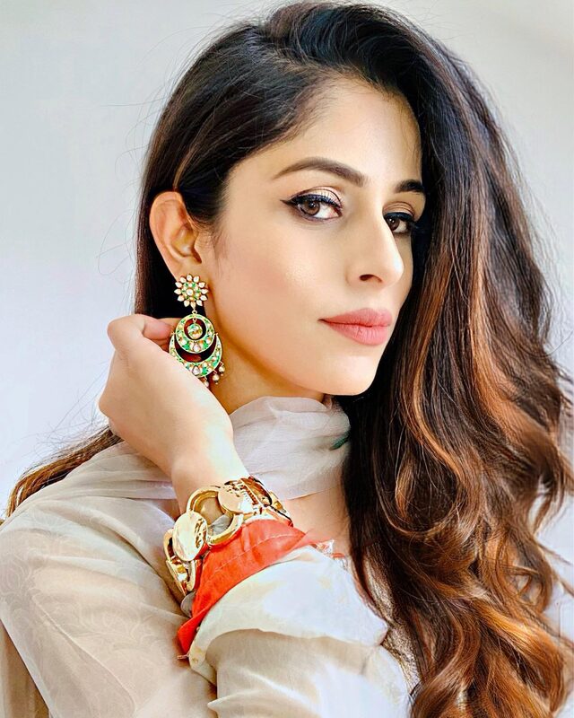 Close-up portrait wearing gold and green kundan-style drop earrings and a gold bangle, with a cream dupatta and a peek of orange fabric at the shoulder