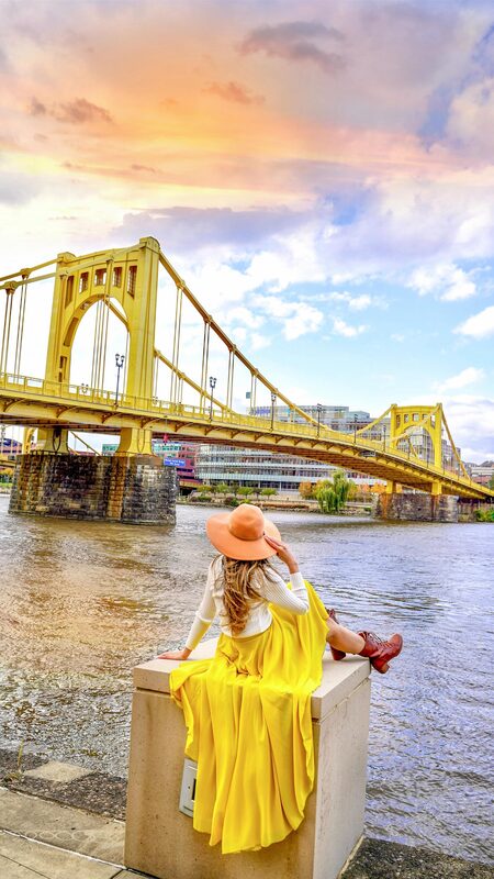 Roopsi Bansal in an orange sun hat, white sweater, and flowing yellow maxi skirt, seated on a stone pedestal beside the Allegheny River with the yellow Rachel Carson Bridge behind her