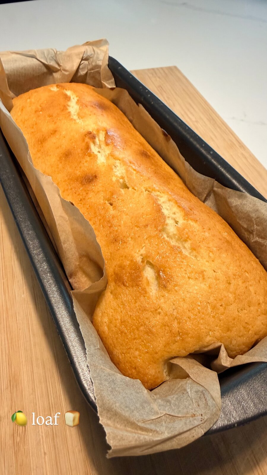 A golden lemon loaf cake, fresh from the oven, cooling in its parchment-lined pan on a wood board