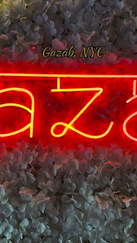 A neon 'Gazab' sign against a wall of flowers at Gazab NYC restaurant