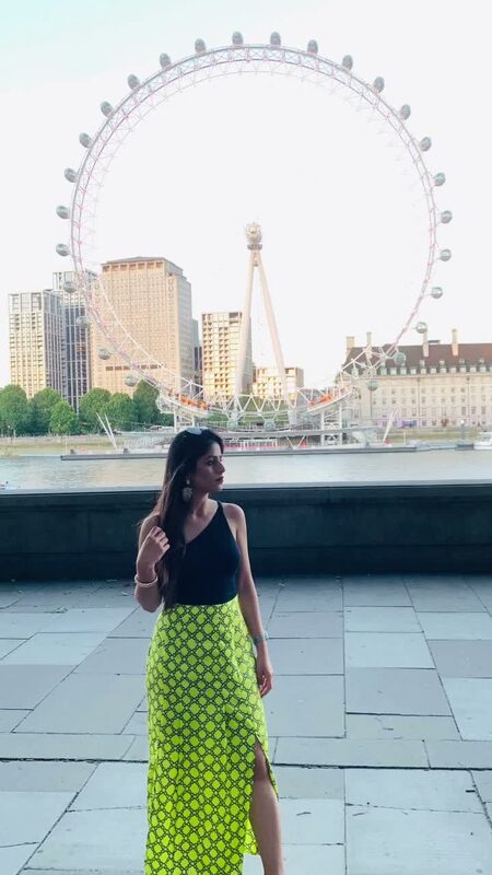 Roopsi Bansal in a black cami top and neon green geometric-print wrap skirt, standing on the South Bank with the London Eye in the background