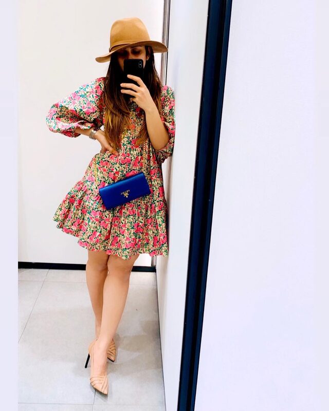 Mirror selfie in a pink and green floral babydoll dress and a wide-brim tan hat, holding a blue Prada wallet, in a plain hotel room