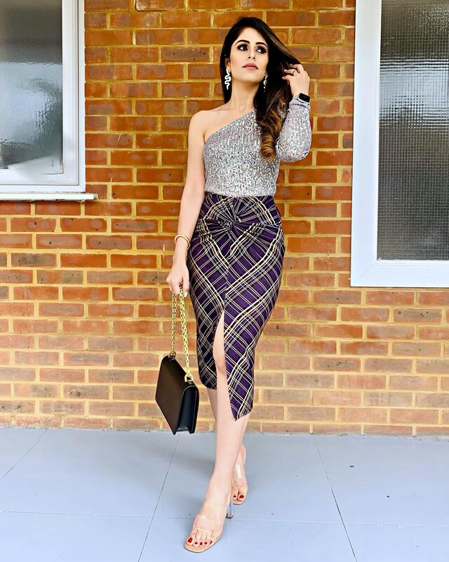 Standing against a brick wall wearing a silver sequin one-shoulder top and a purple plaid wrap skirt with a front slit, snake-drop earrings, holding a black clutch