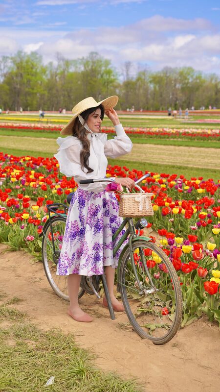 Roopsi Bansal in a white ruffle blouse and purple floral skirt with a straw hat, leaning on a vintage bicycle in a rainbow tulip field