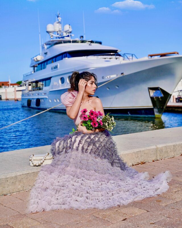Roopsi Bansal in a lavender ruffled tulle skirt and blush puff-sleeve top, holding a bouquet of pink anemones at a marina in West Palm Beach with a superyacht docked behind her