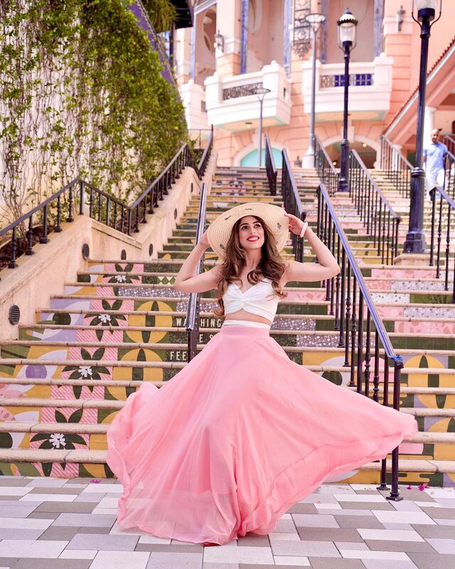 Roopsi Bansal in a white crop top and flowing pink maxi skirt with a straw hat, twirling on a colourful mosaic-tiled staircase at Rosemary Square, West Palm Beach