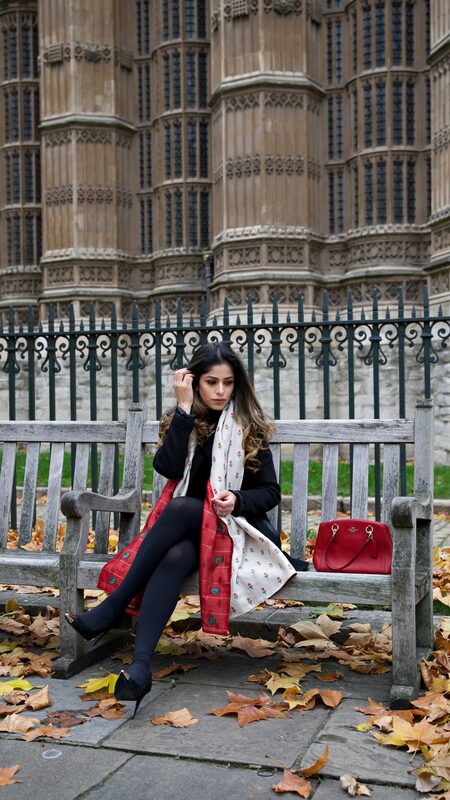 Roopsi Bansal sitting on a wooden bench draped in a red and white floral scarf, a red Coach handbag beside her, in front of a Gothic stone building with autumn leaves on the ground