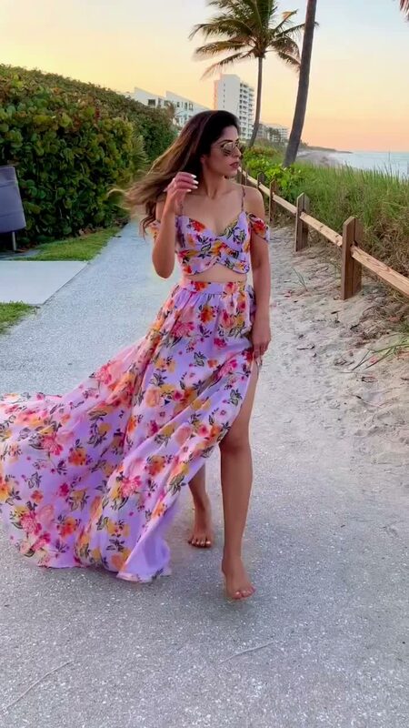 Roopsi Bansal in a lilac floral print cold-shoulder crop top and matching split-thigh maxi skirt from SHEIN, walking a beachside boardwalk at sunset with palm trees and the ocean behind her