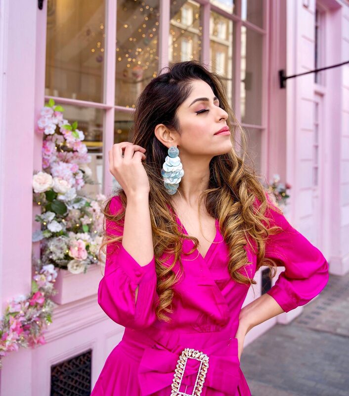 Roopsi Bansal in a hot-pink wrap dress with a crystal buckle belt and statement drop earrings, standing outside a pink storefront decorated with fresh flowers