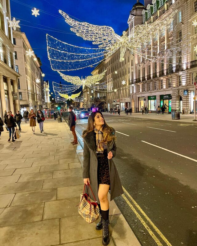 Roopsi Bansal in a grey coat with a Burberry check scarf and tote and black lace-up boots, standing on a London street strung with glittering angel-shaped light installations at night
