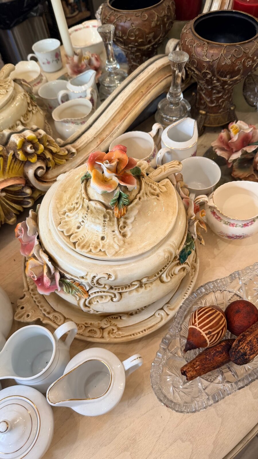 A tabletop crowded with antique porcelain teacups, gilded vases, crystal candlesticks, and a flower-topped tureen in a curio shop