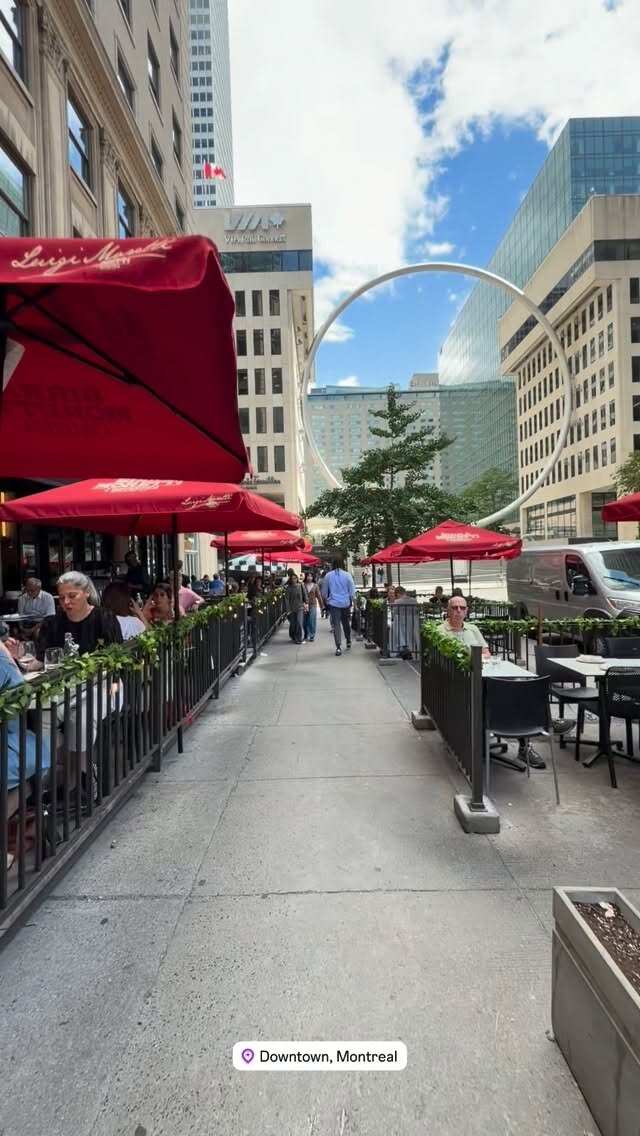 A sidewalk lined with red umbrella-shaded restaurant patios in downtown Montreal, with a large arched sculpture overhead