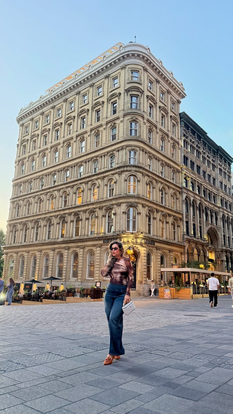 Roopsi Bansal walking across Place D'Armes in Old Montreal in a tie-dye top and flared jeans, with the ornate Hotel Place D'Armes facade behind her