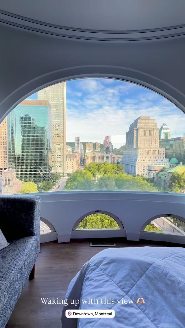 A downtown Montreal skyline seen through a large arched hotel window, with a bed and sofa in the foreground