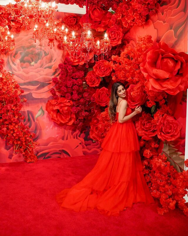 Roopsi Bansal in a red tiered ball gown from Lunamis, standing in a room filled with red roses and red chandeliers at The Floral Escape