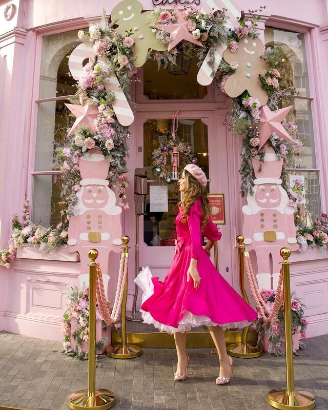 Roopsi Bansal in a hot pink puff-sleeve dress with a white ruffled underskirt and a pink beret, twirling outside a pink shopfront decorated with gingerbread-man cutouts and floral wreaths