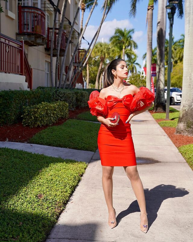 Roopsi Bansal in a red off-shoulder bandage dress with red organza ruffle sleeves and a heart bow, holding a heart-shaped red clutch, on a palm-lined street