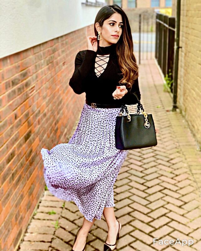 Walking down a brick alley in a black lace-up top and a lavender polka-dot pleated skirt with a Gucci logo belt, carrying a black Michael Kors tote