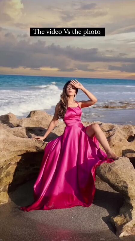 Roopsi Bansal in a fuchsia satin gown seated on beach rocks at dusk, waves breaking behind her
