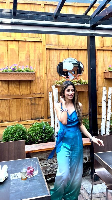 Roopsi Bansal in a blue-to-teal ombré draped cami jumpsuit, standing on a wooden-fenced restaurant patio