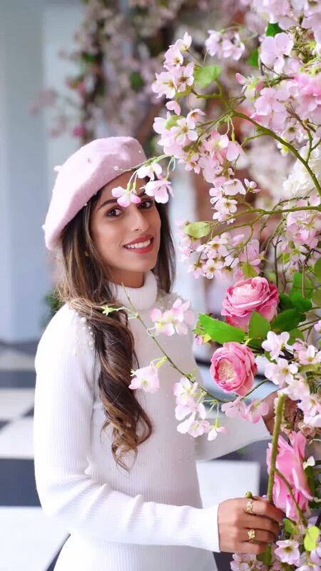 Roopsi Bansal in a white ribbed turtleneck sweater dress and a pink beret, smiling beside a display of pink cherry blossom branches