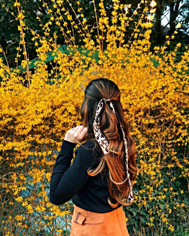 Back-of-head view of a leopard-print ribbon tied into half-up hair, wearing a black turtleneck and orange skirt, standing before a bush covered in bright yellow flowers