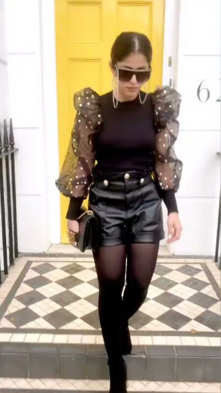 Roopsi Bansal in a black top with sheer polka-dot puff sleeves and leather shorts, walking down steps in front of a yellow door