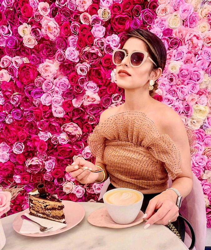 Roopsi Bansal in a sequined brown top and sunglasses, seated at a table with cake and coffee in front of a wall of pink roses at Cafe Chloe in Prague