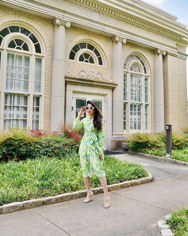 Roopsi Bansal in a green and white ikat-print silk wrap dress from MOTF, white sunglasses pushed down her nose, standing in front of the Civic Hall building
