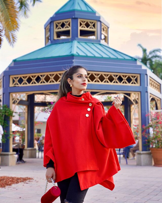 Roopsi Bansal in a red asymmetric hem button wrap cape coat from Chicwish, standing at a blue-and-gold gazebo