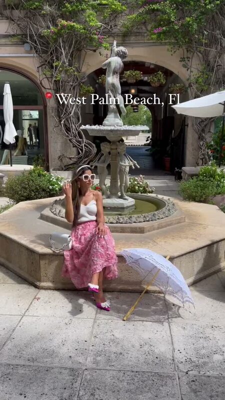 Roopsi Bansal in a pink tulle skirt and white top seated at a fountain courtyard in West Palm Beach, holding a white lace parasol